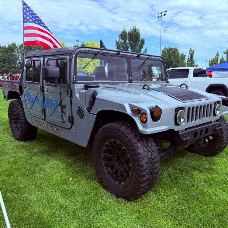 Grey Hummer H1 truck with Blue Zero branding parked on grass under an American flag
